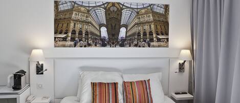 A hotel room with a large bed, two pillows, a nightstand with a lamp, a phone, and a framed picture of a busy street scene on the wall.
