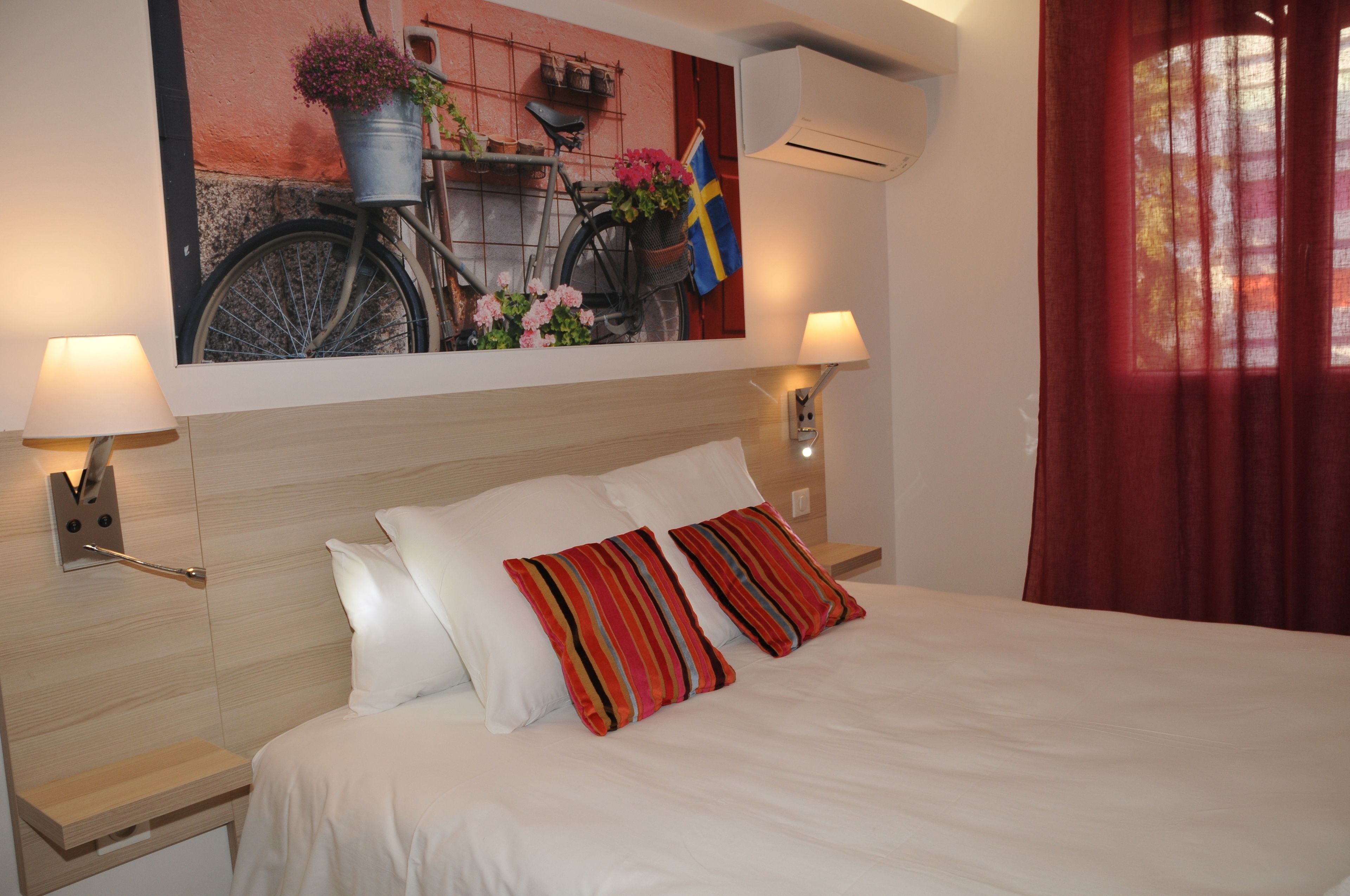 A hotel room with a bed, two pillows, a nightstand with a lamp, a wall-mounted air conditioner, and a painting of a bicycle with flowers.