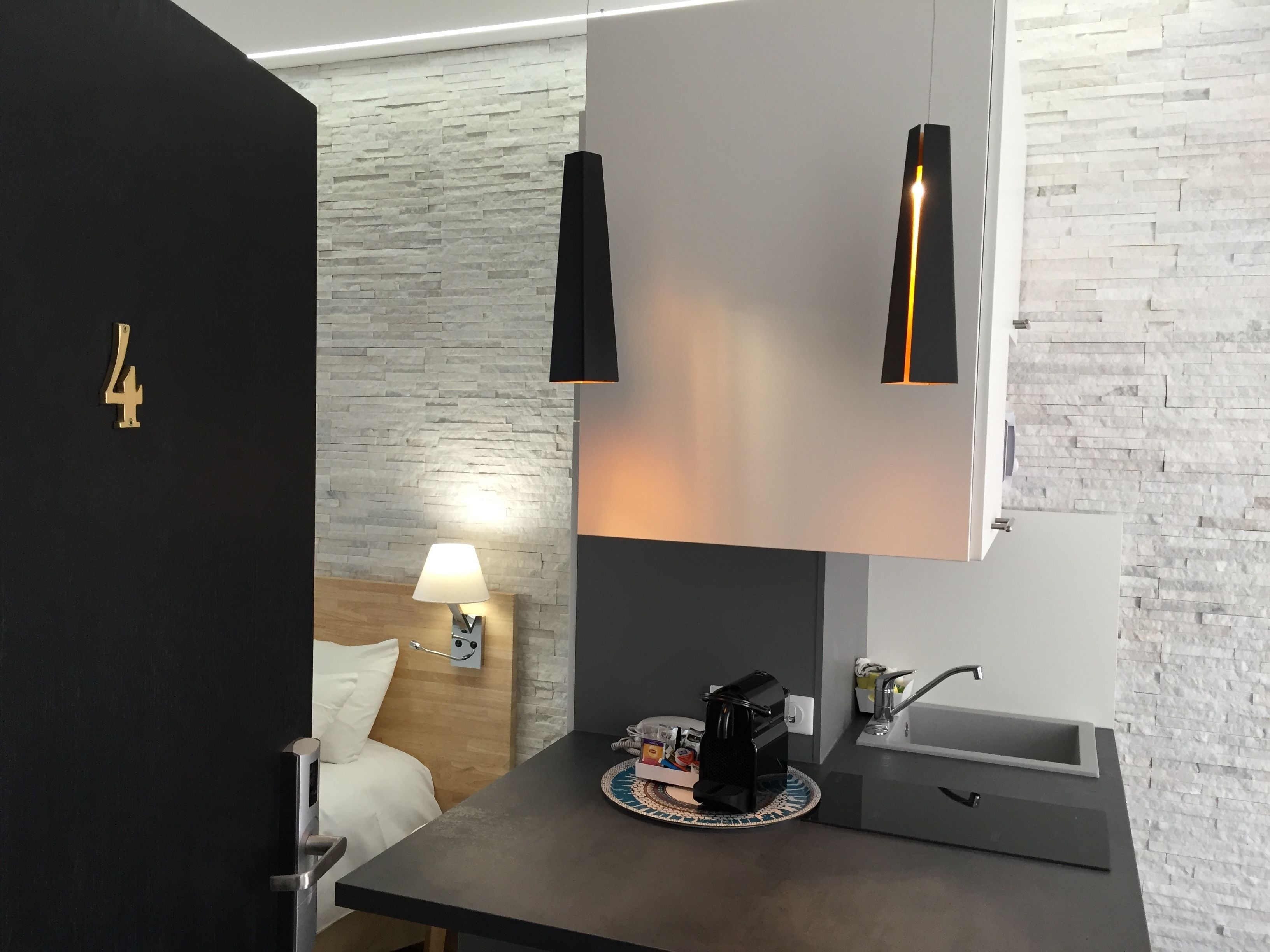 Ein modernes Hotelzimmer mit einer Wand im Naturstein-Look, einem Bett mit weißer Bettwäsche, einem Nachttisch mit Lampe, einer Kaffeemaschine und einem kleinen Waschbecken.