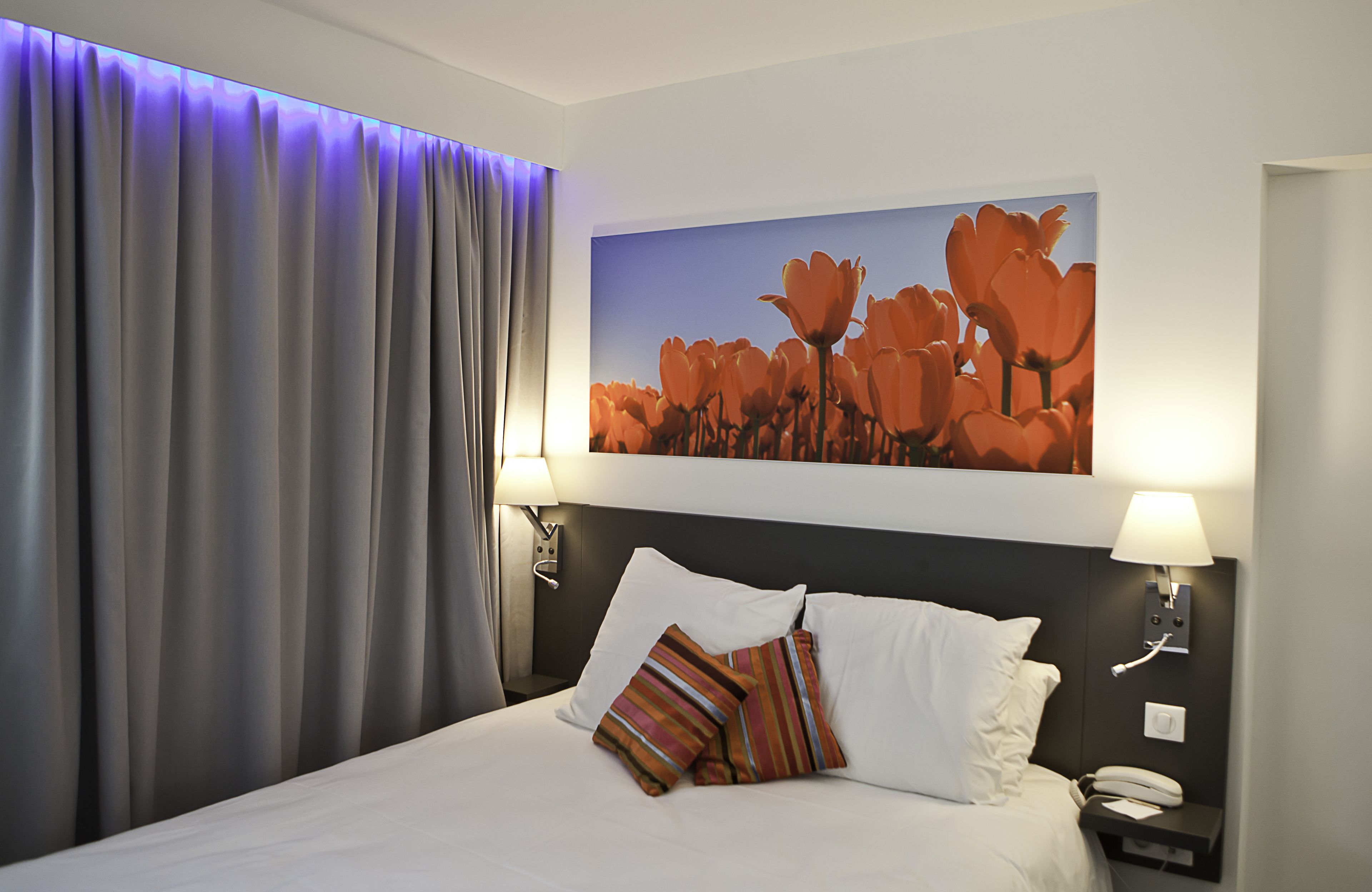 A hotel room with a large bed, two bedside lamps, a nightstand with a phone, and a painting of flowers on the wall.