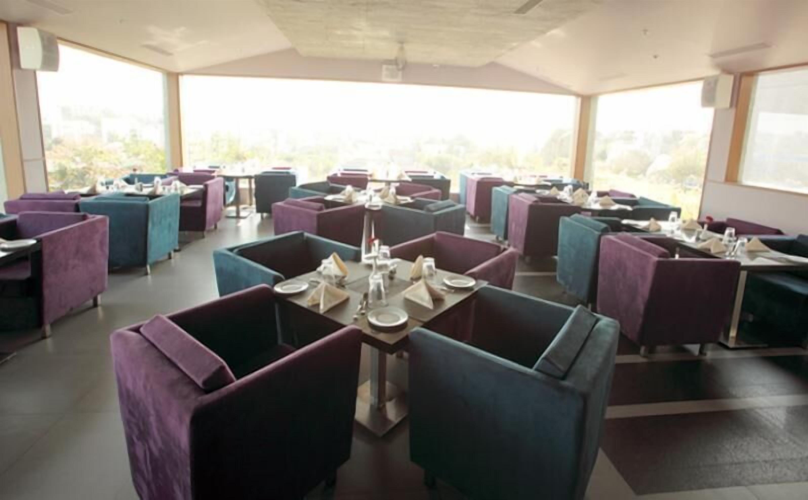 A restaurant with tables and chairs arranged in a grid pattern, large windows allowing natural light, and a view of greenery outside.