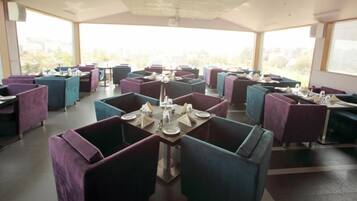 A restaurant with tables and chairs arranged in a grid pattern, large windows allowing natural light, and a view of greenery outside.