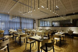 En modern restaurang med flera bord och stolar, ett högt tak med hängande lampor och stora fönster med gardiner.