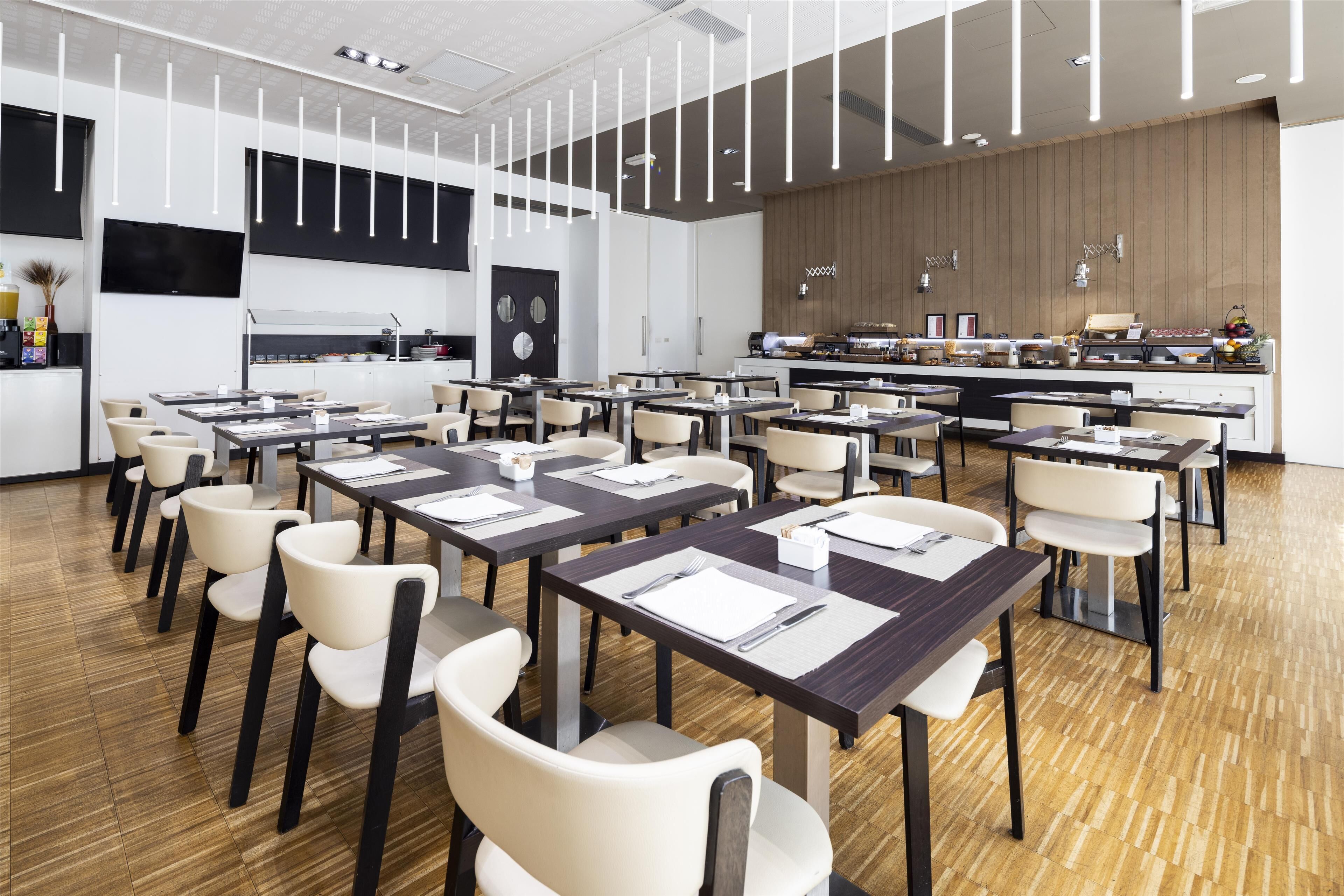 A modern dining area with multiple tables and chairs, a kitchen in the background, and a wooden wall.
