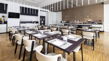 A modern dining area with multiple tables and chairs, a kitchen in the background, and a wooden wall.