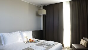 Un cuarto de hotel con una cama bien hecha, una bandeja con alimentos para el desayuno, una mesita de noche con lámpara y vistas al exterior a través de cortinas amplias.