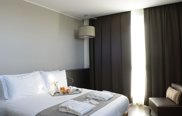Une chambre d’hôtel avec un lit parfaitement fait, un plateau de petit-déjeuner, une table de chevet avec une lampe et une vue sur l’extérieur à travers de grandes rideaux.