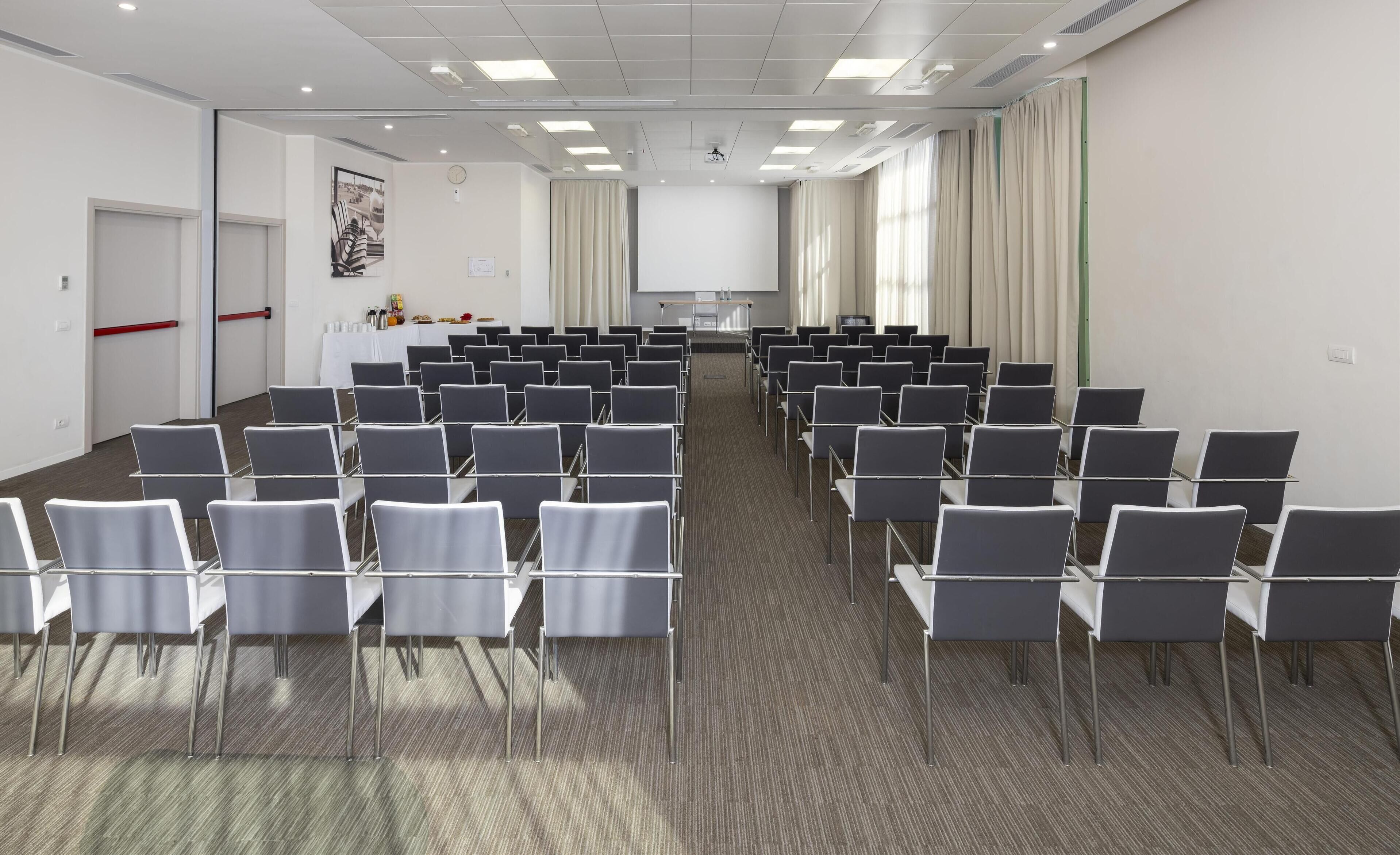 A conference room with rows of chairs, a projector screen, and a table with refreshments.
