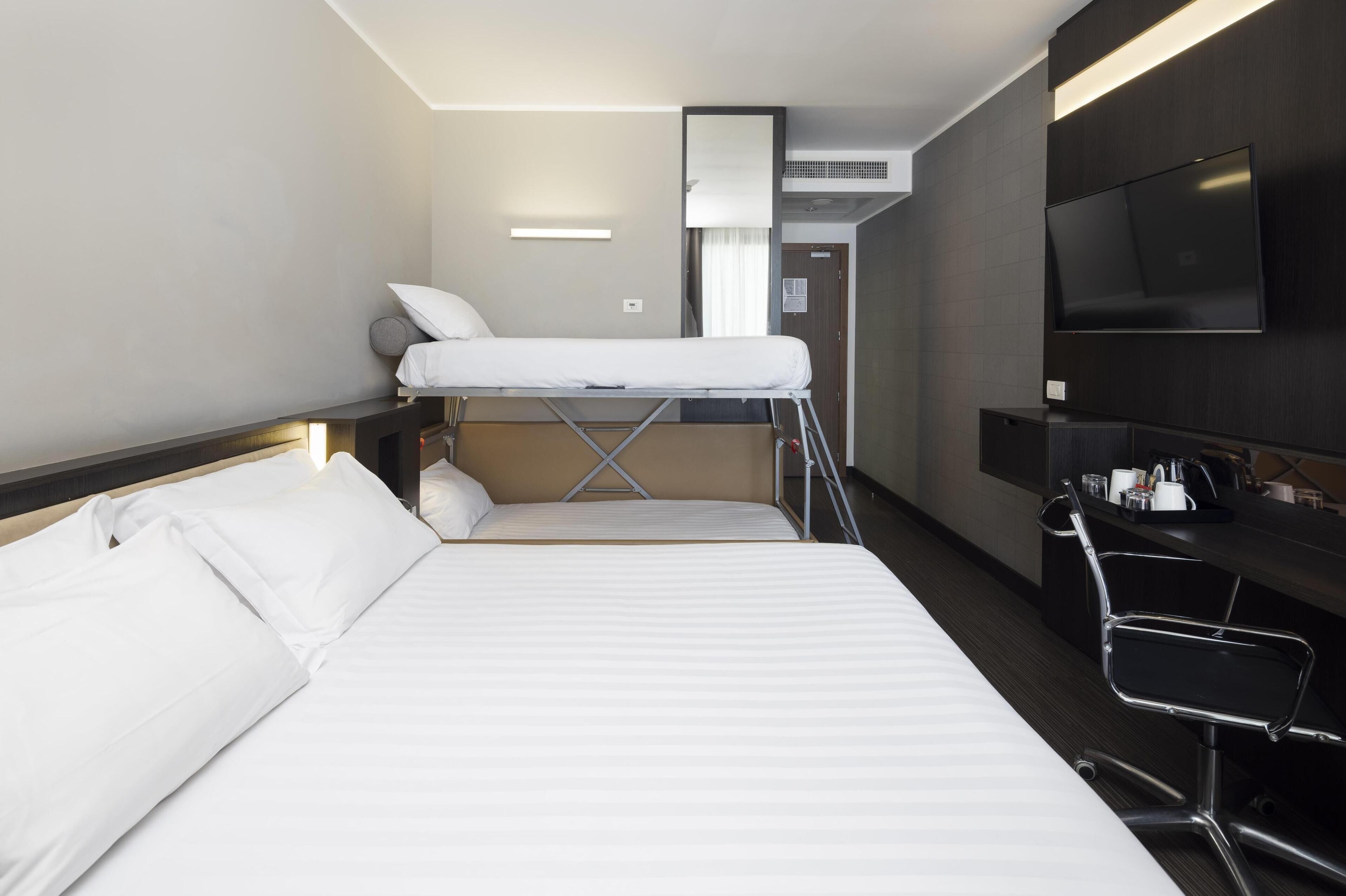 A hotel room with a bunk bed, a desk with a chair, a flat-screen TV, and a minibar.