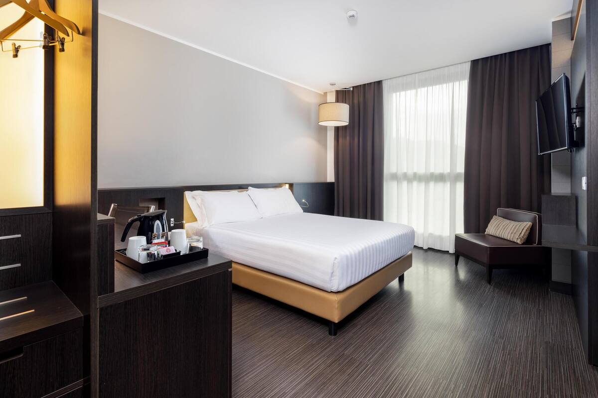 Een moderne hotelkamer met een groot bed, een nachtkastje met koffiezetapparaat, een televisie en een zithoek met een comfortabele bank.