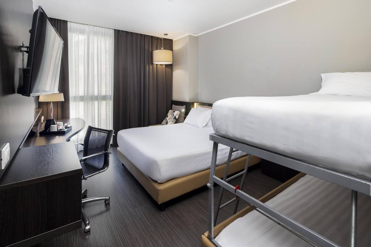 Een hotelkamer met een bureau, twee bedden, een stoel, een televisie en een raam met gordijnen.