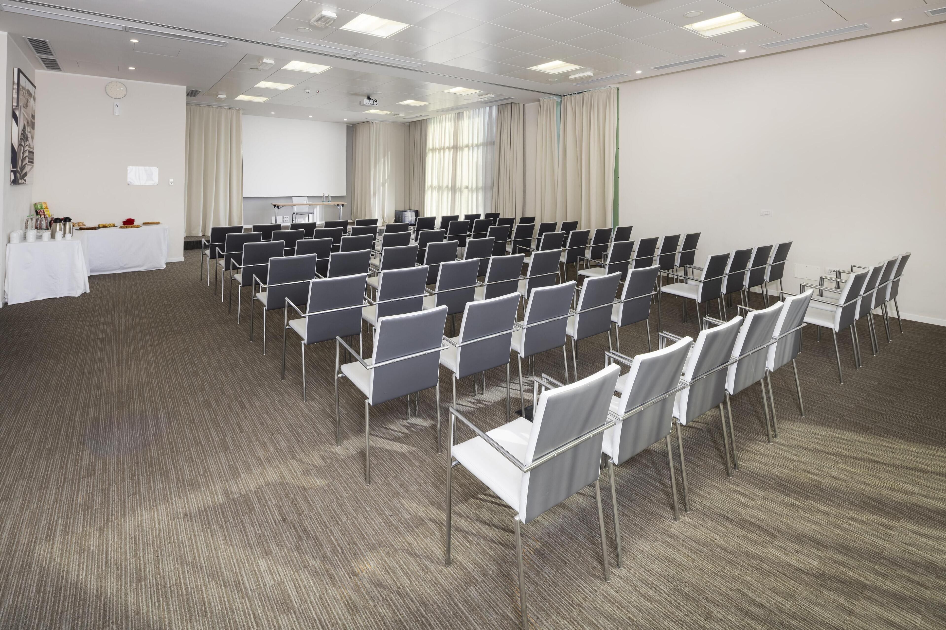 Un salón de conferencias con filas de sillas, una pantalla de proyector y una mesa con refrigerios.