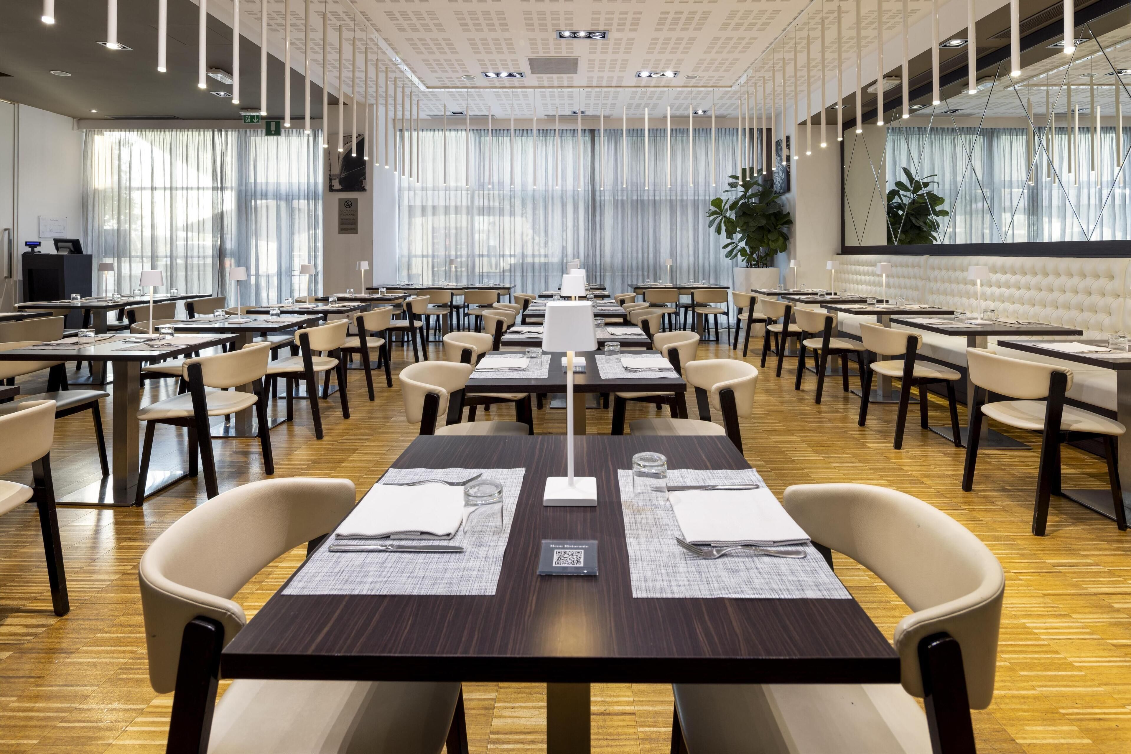 Un'ampia sala da pranzo con diversi tavoli e sedie, grandi finestre con tende leggere e un moderno design del soffitto.