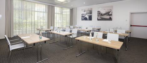 A conference room with multiple tables and chairs, large windows with curtains, and two black and white car photographs on the wall.
