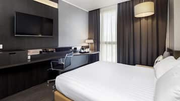 Ein Hotelzimmer mit einem Bett, einem Schreibtisch mit Stuhl, einem Flachbildfernseher, einer Lampe und einem großen Fenster mit Vorhängen.