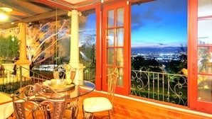 A dining area with a table and chairs, a decorative column, and a view of a cityscape at dusk. - Ringle Resort & Spa (Escazu)
