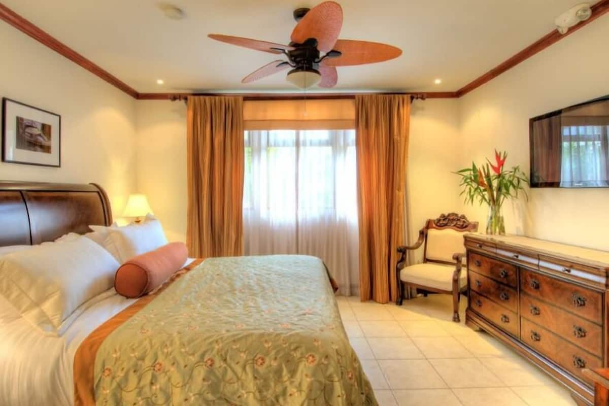 A hotel room with a bed, a dresser, a chair, a ceiling fan, and a window with curtains.