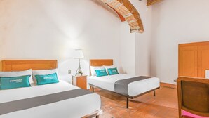 A hotel room with two beds, each with a wooden headboard and teal pillows. There is a wooden chair with a pink cushion, a nightstand with a lamp, and a brick wall arch. - Ayenda Meson de San Sebastian (Puebla)