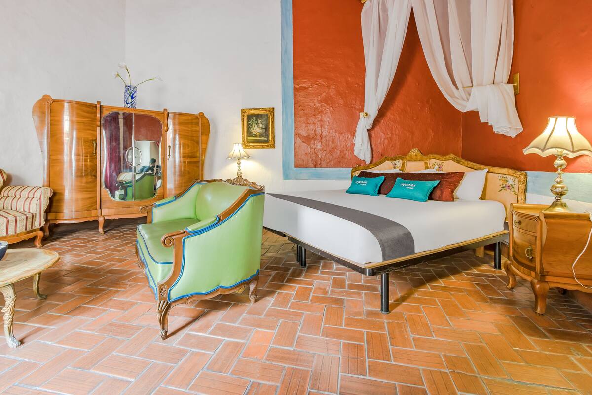 Een hotelkamer met een bed, een groene fauteuil, een houten garderobe en een spiegel.