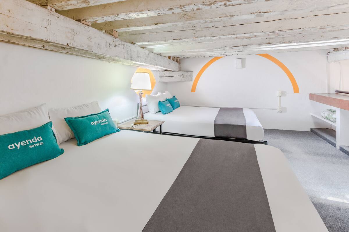 Een moderne hotelkamer met een bed, een bureau en een stoel. Er staat een lamp op het bureau en een kussen met de naam van het hotel.