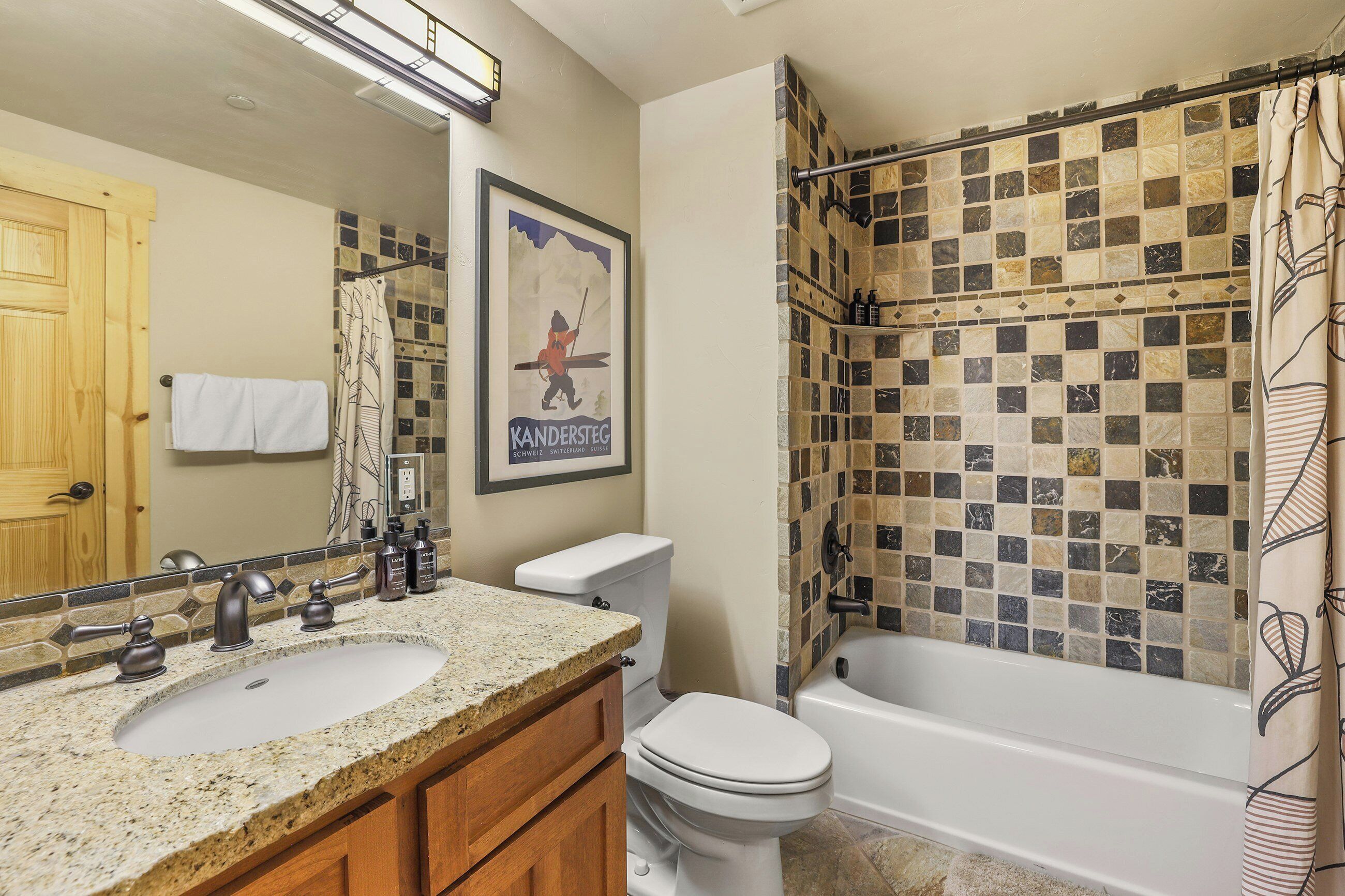 A bathroom with a granite countertop sink, a toilet, and a bathtub with mosaic tile walls.