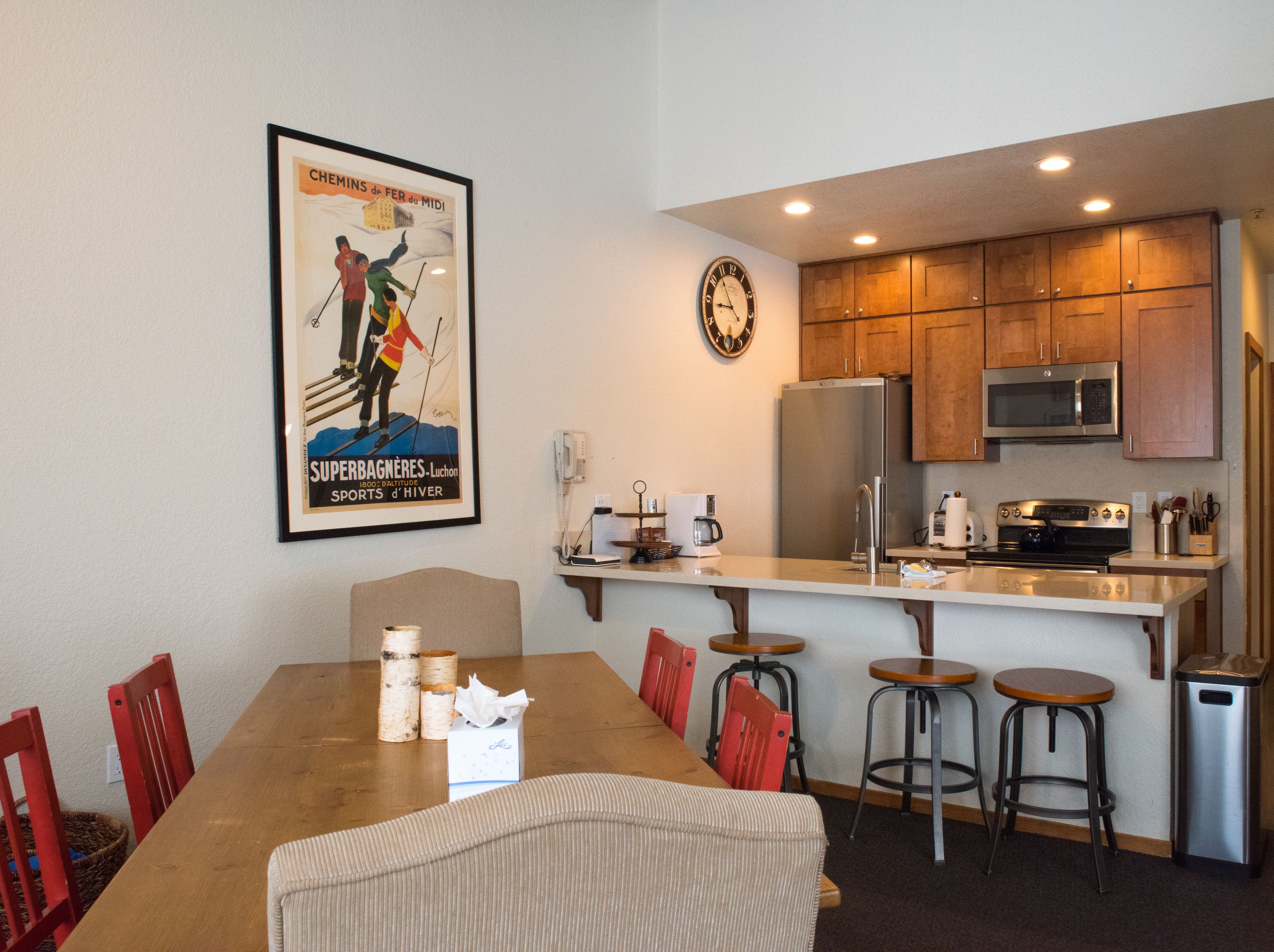 Una cocina moderna con armarios de madera, electrodomésticos de acero inoxidable y una zona de comedor con mesa de madera y sillas rojas.
