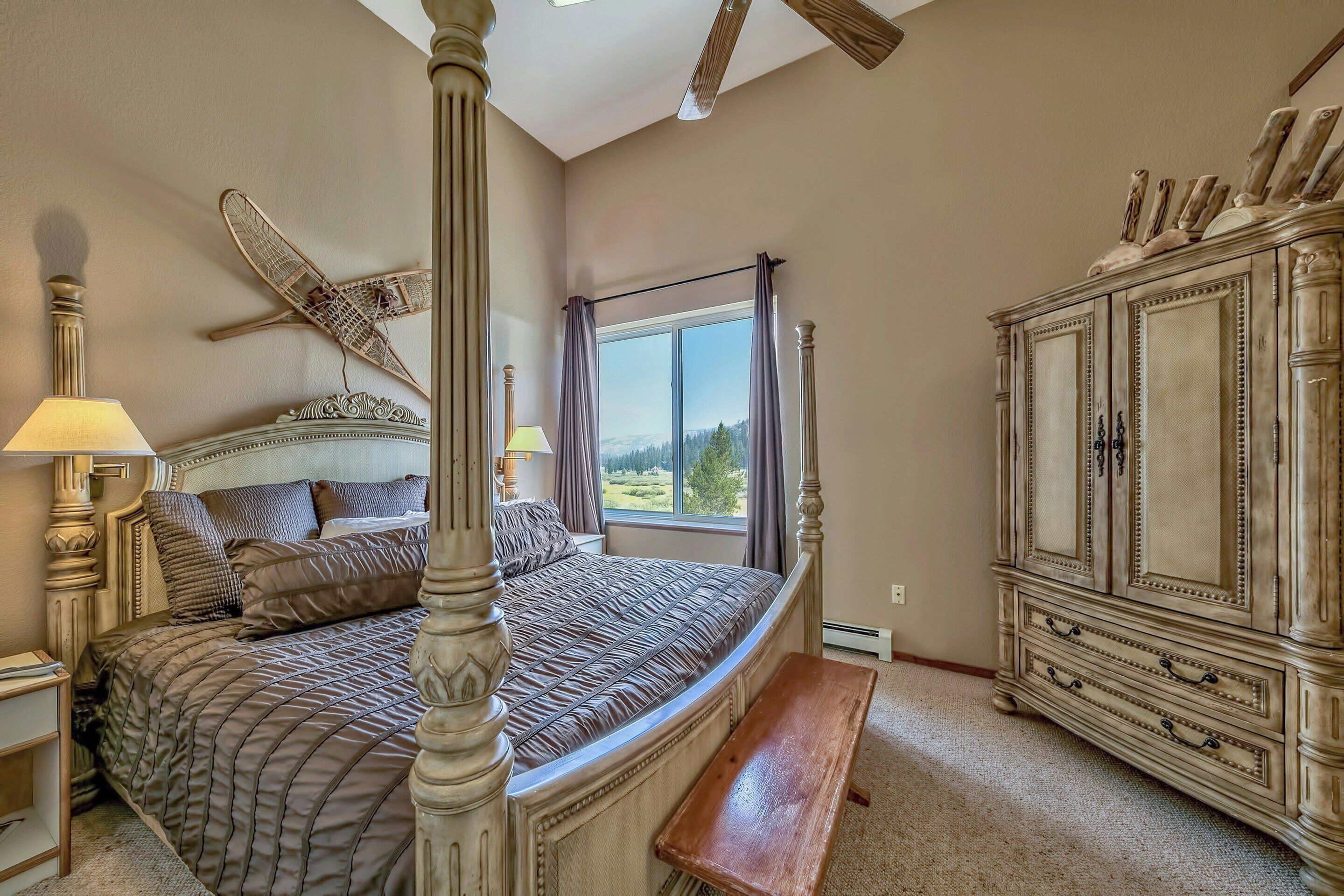 A bedroom with a four-poster bed, a large window with curtains, a wooden bench, and a classic dresser.