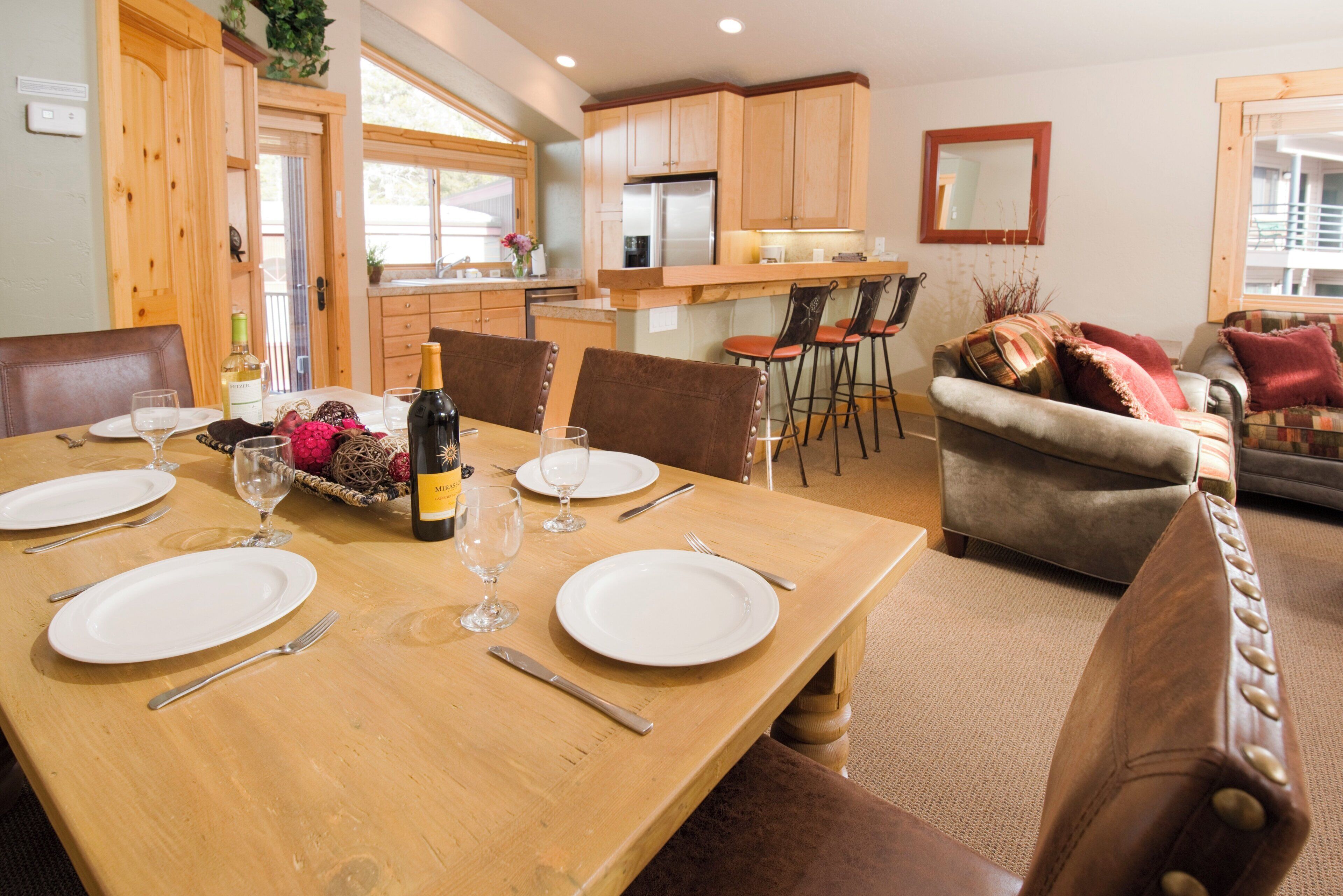 Un comedor con una mesa de madera dispuesta para cuatro, una cocina al fondo y una sala de estar con sofá y sillones.