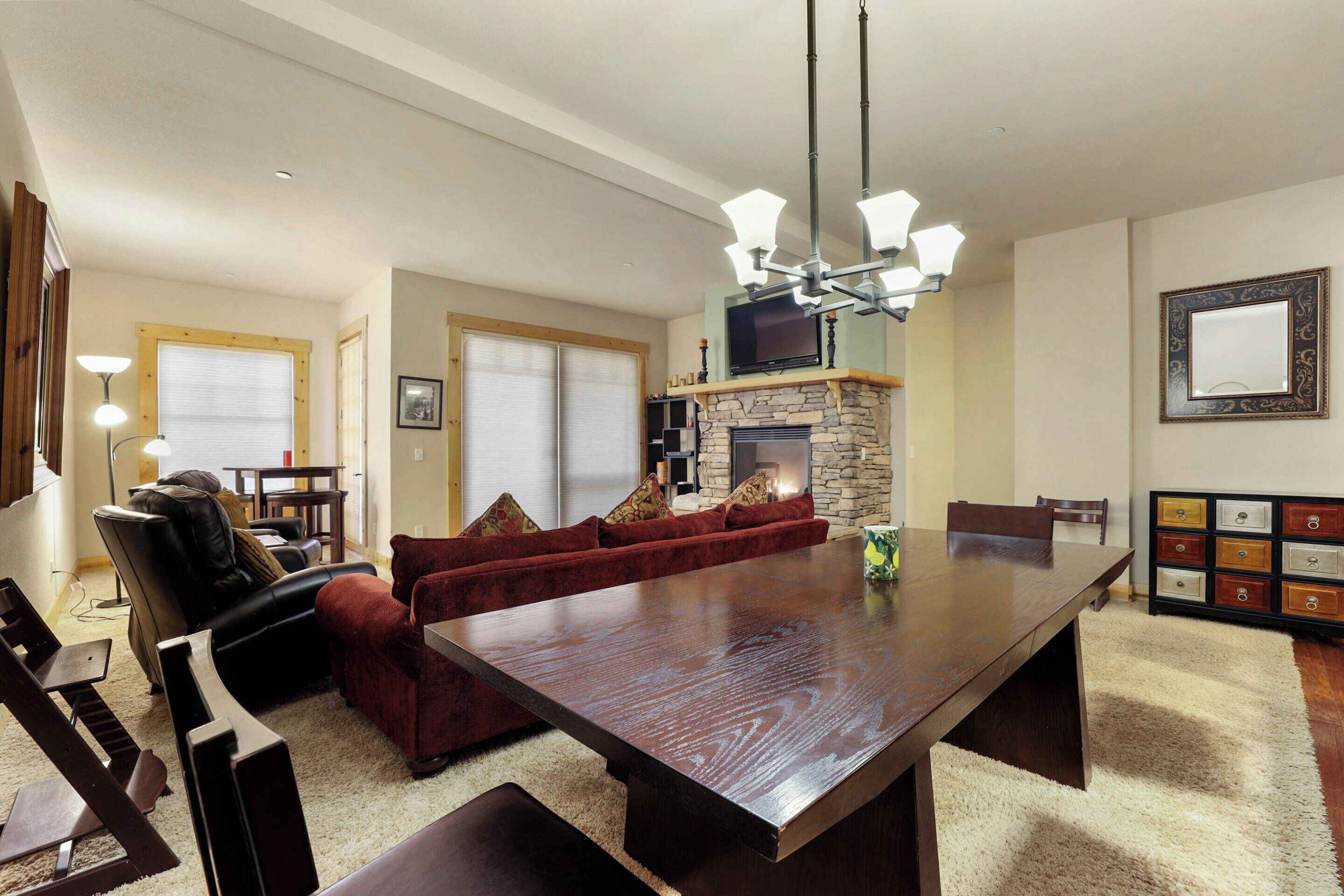 A spacious living room with a large wooden dining table, a red velvet sofa, and a stone fireplace.