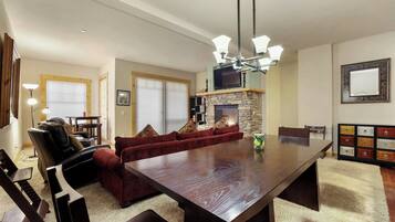 A spacious living room with a large wooden dining table, a red velvet sofa, and a stone fireplace.