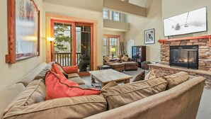 A spacious living area with a stone fireplace, comfortable seating, and a large window overlooking a deck. - Kirkwood Mountain Resort by Vail Resorts (Kirkwood)
