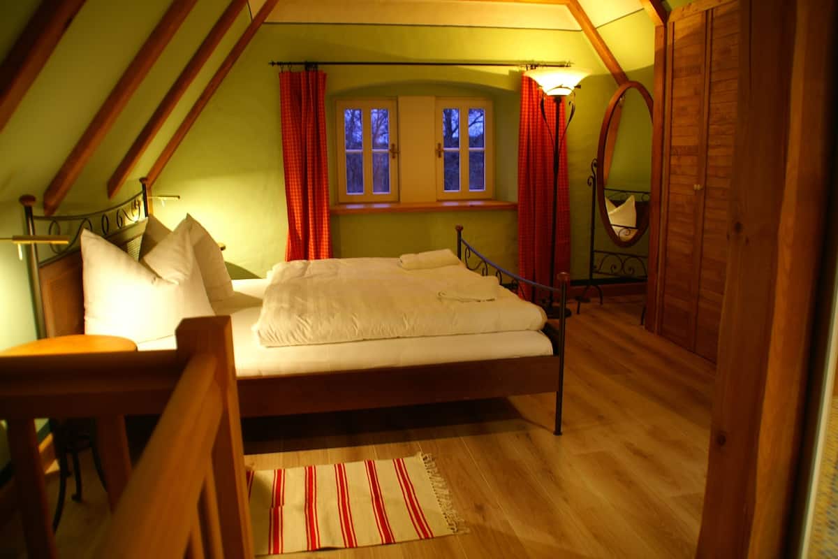A bedroom with a wooden bed, a desk, a chair, a window with curtains, and a mirror.