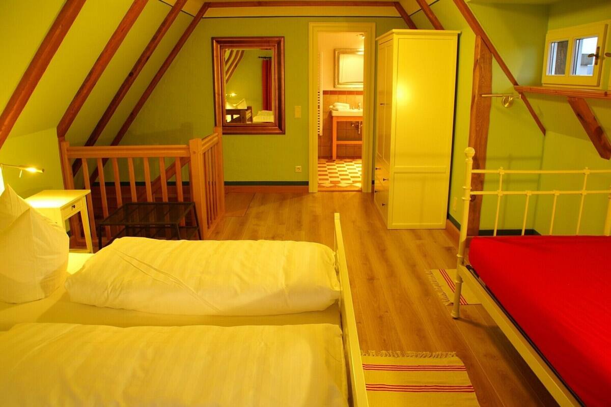 A bedroom with a loft bed, a small table with a lamp, a wooden chair, and a red bedspread.