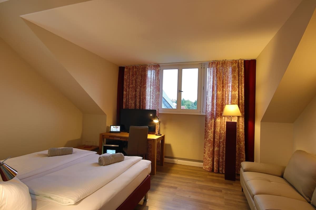 A hotel room with a bed, a desk with a computer, a chair, a sofa, a window with curtains, and a lamp.