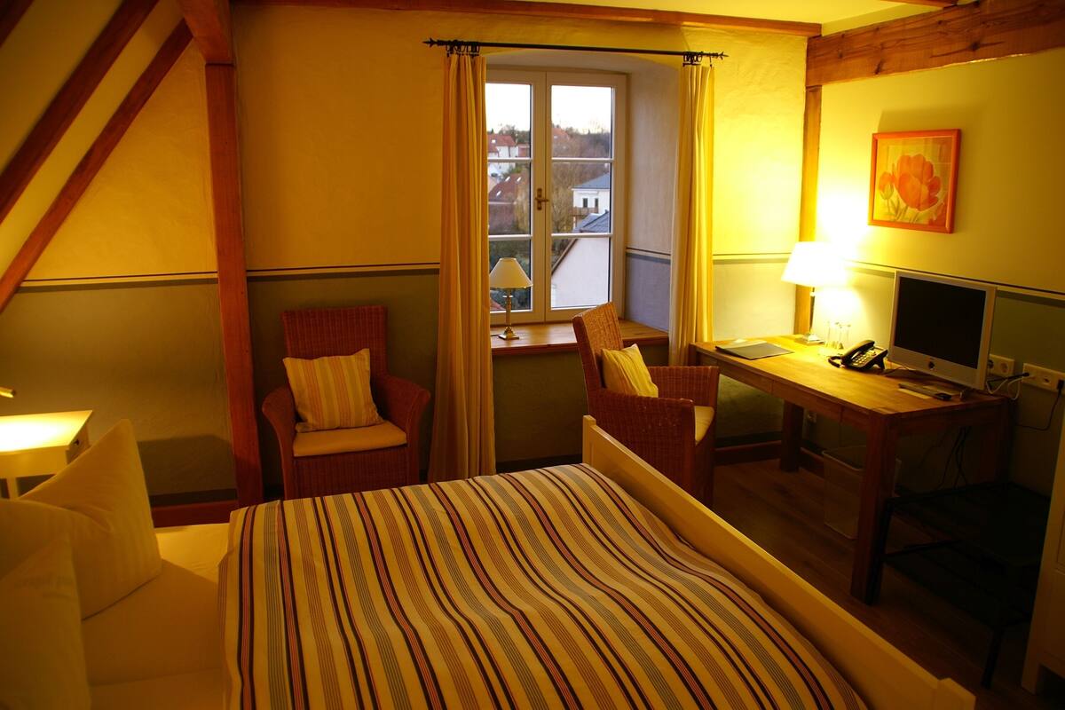 A hotel room with a bed, two chairs, a desk with a computer, and a window with curtains.