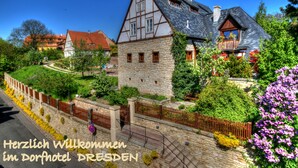 A traditional half-timbered building with a stone base and a sloped roof, surrounded by lush greenery and a stone wall with a gate.