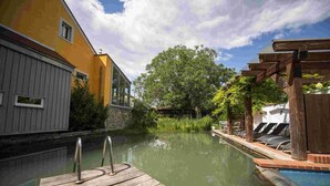 A wooden dock leading into a calm water body, surrounded by lush greenery and a yellow building.
