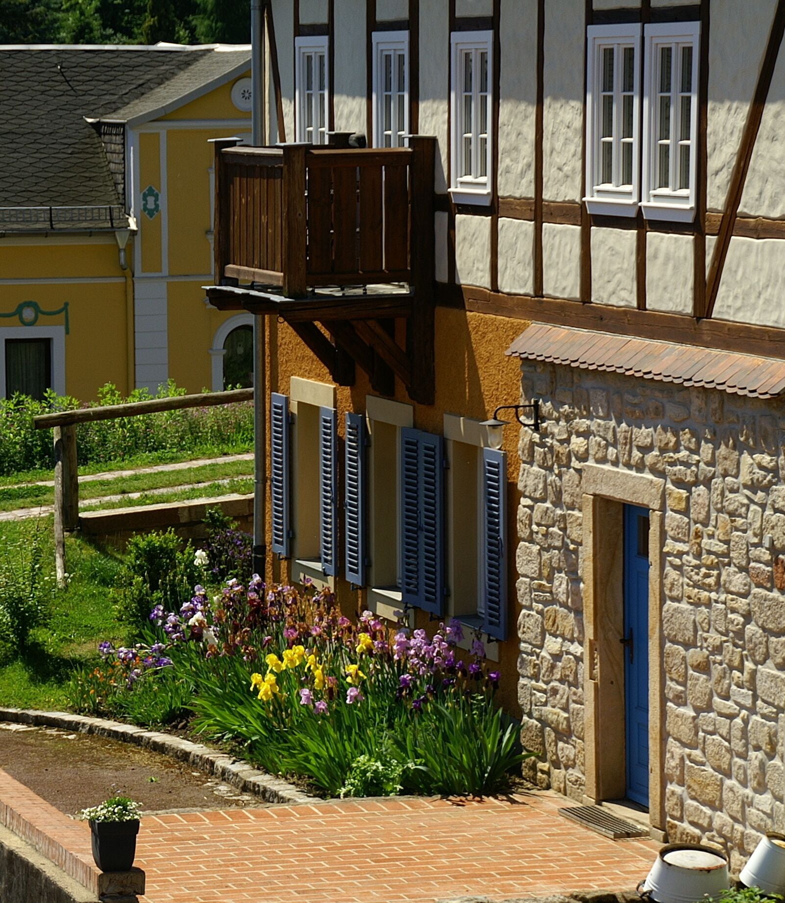 A traditional half-timbered building with a stone facade, a wooden balcony, and a flower garden in front.