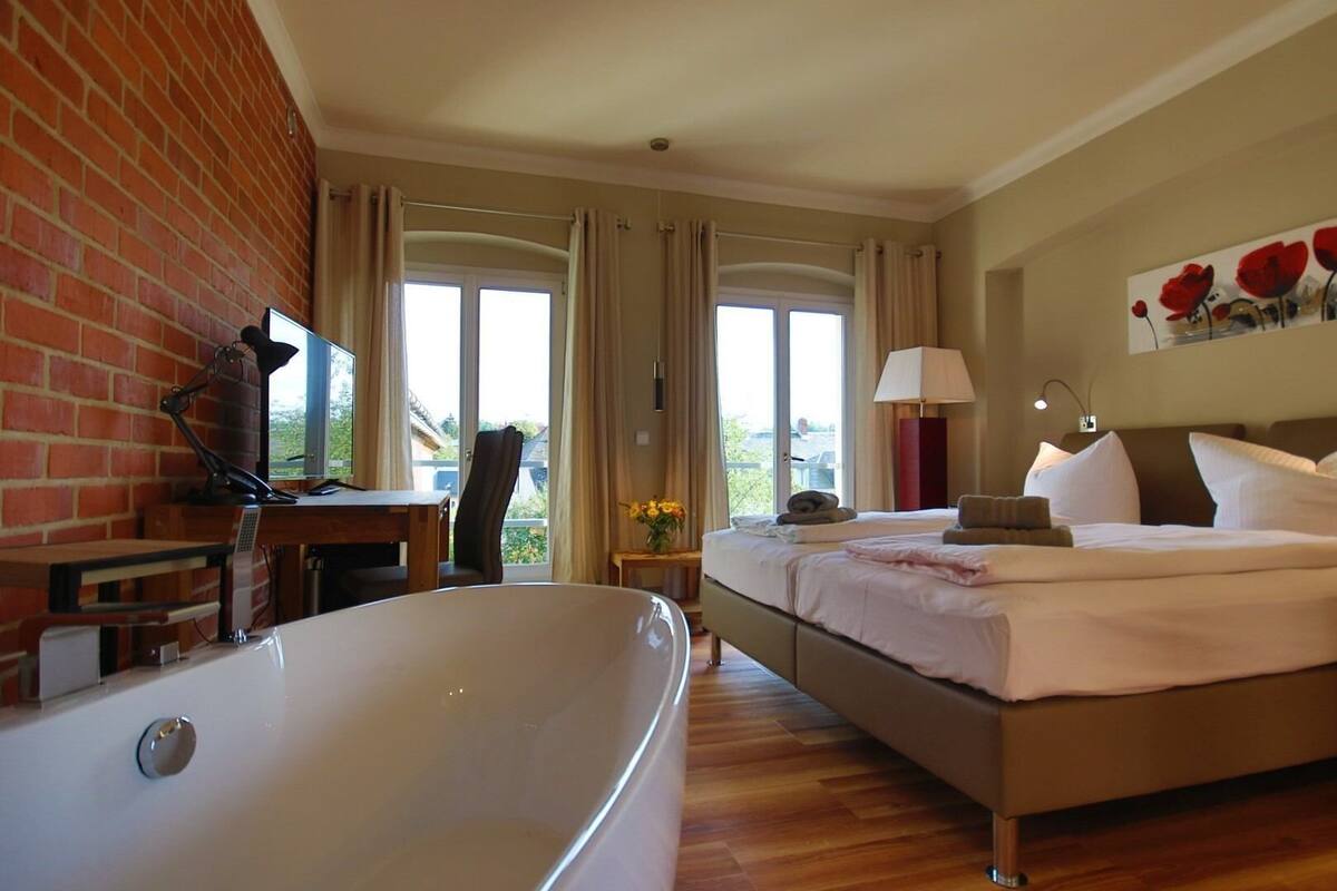 A hotel room with a large bathtub, a bed with two pillows, a desk with a chair, and a window with curtains.