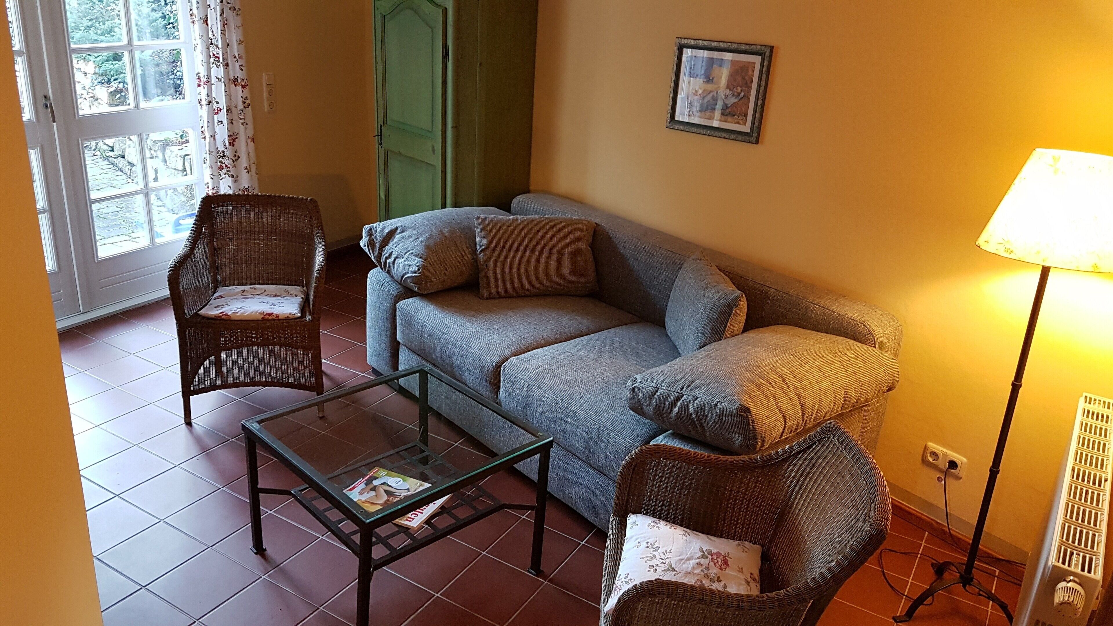 Una sala de estar con un sofá, dos sillas de mimbre, una mesa de centro de vidrio, una lámpara y una puerta que da al exterior.