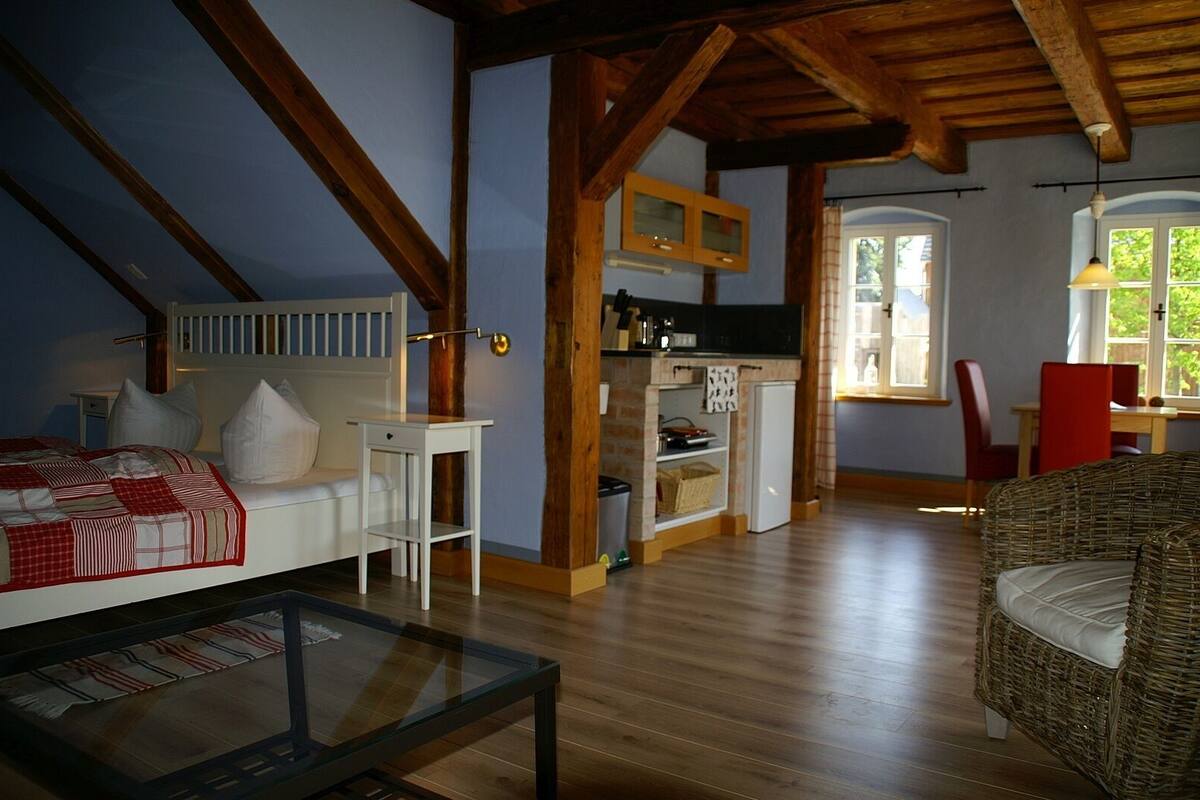 A cozy room with a bed, a glass-top table, a wicker chair, a kitchen area, and a dining table with chairs.