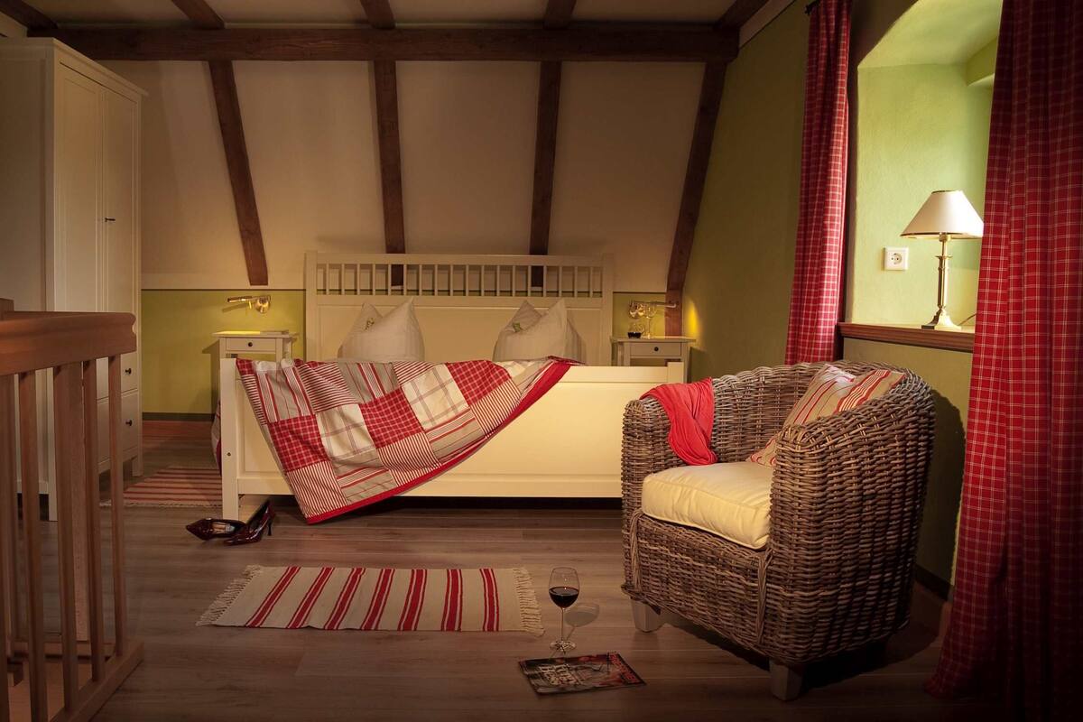 A bedroom with a bed, a wicker chair, a nightstand, a lamp, and a window with curtains.
