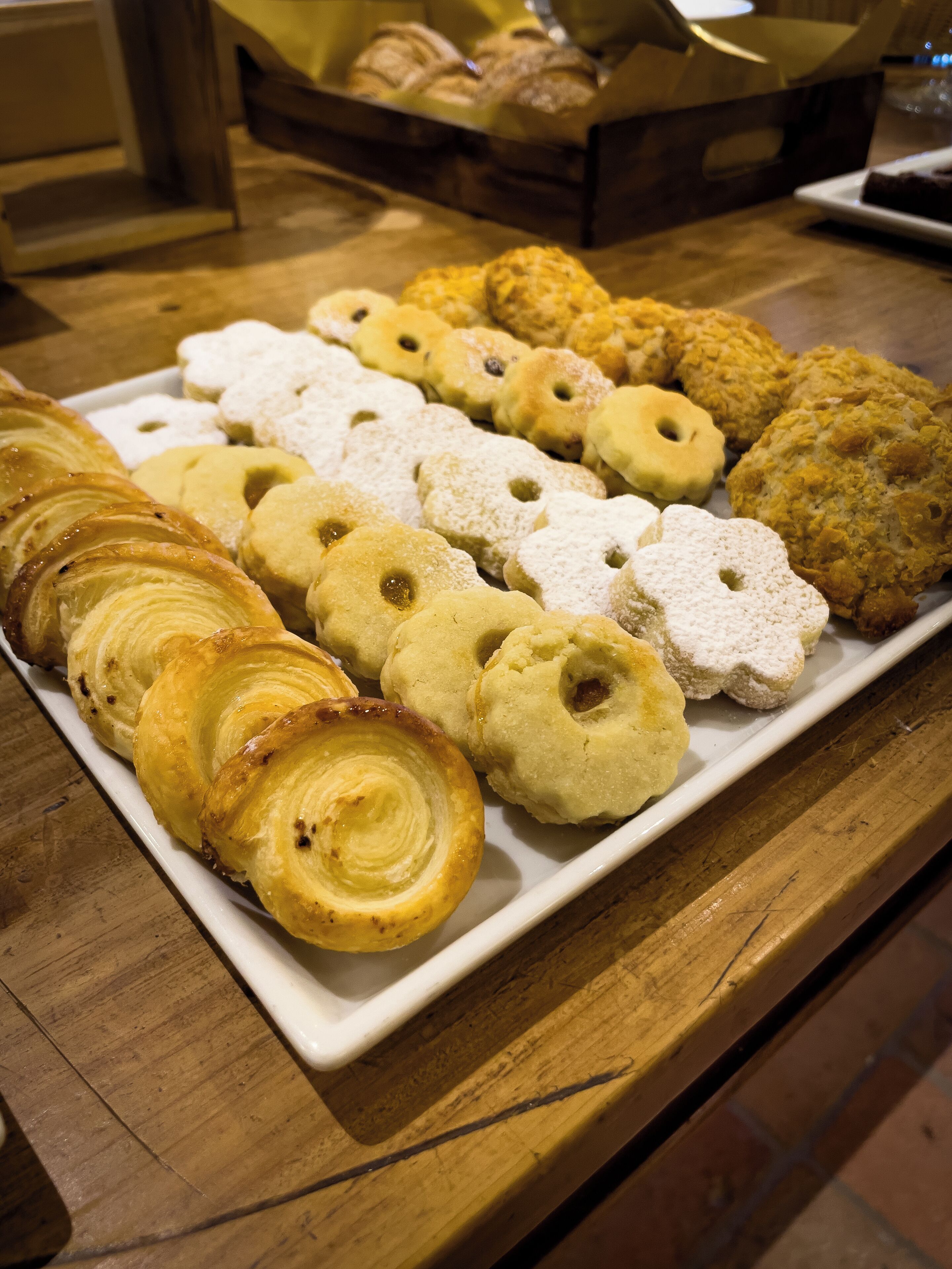Una variedad de pasteles en un plato blanco, incluyendo croissants y galletas, sobre una mesa de madera.