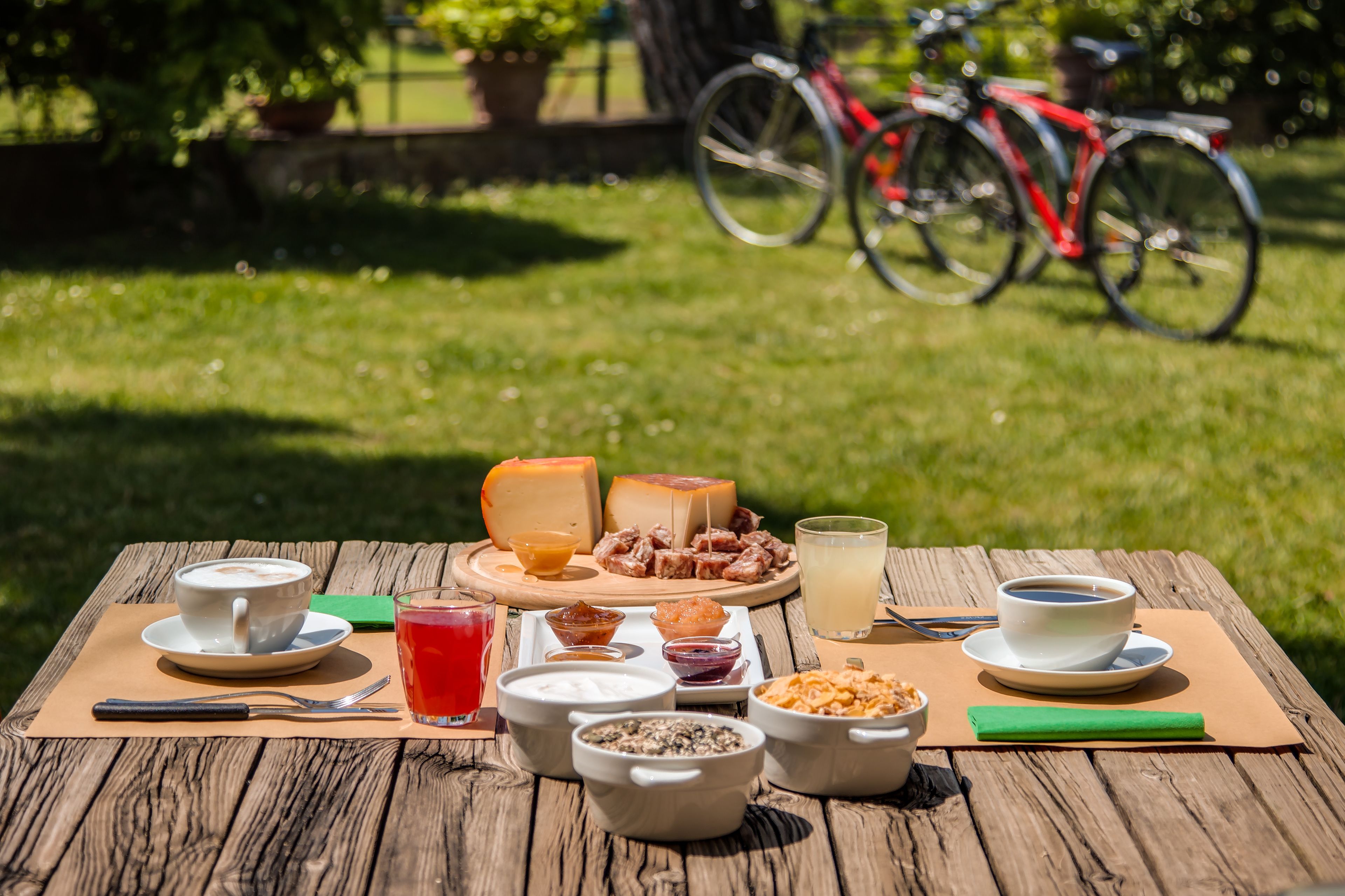 Un coin pique-nique avec de la nourriture et des boissons sur une table en bois, des vélos en arrière-plan et une zone herbeuse.