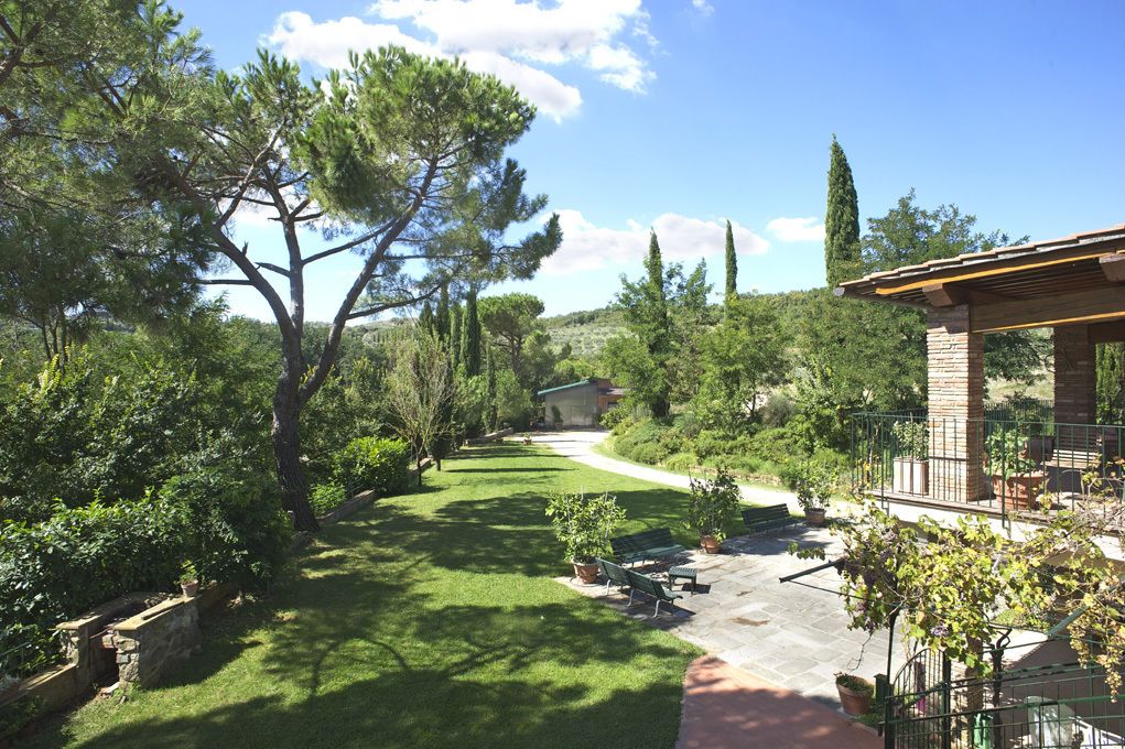 Un jardin avec un chemin menant à un bâtiment. On y trouve des arbres, un banc et une pergola.