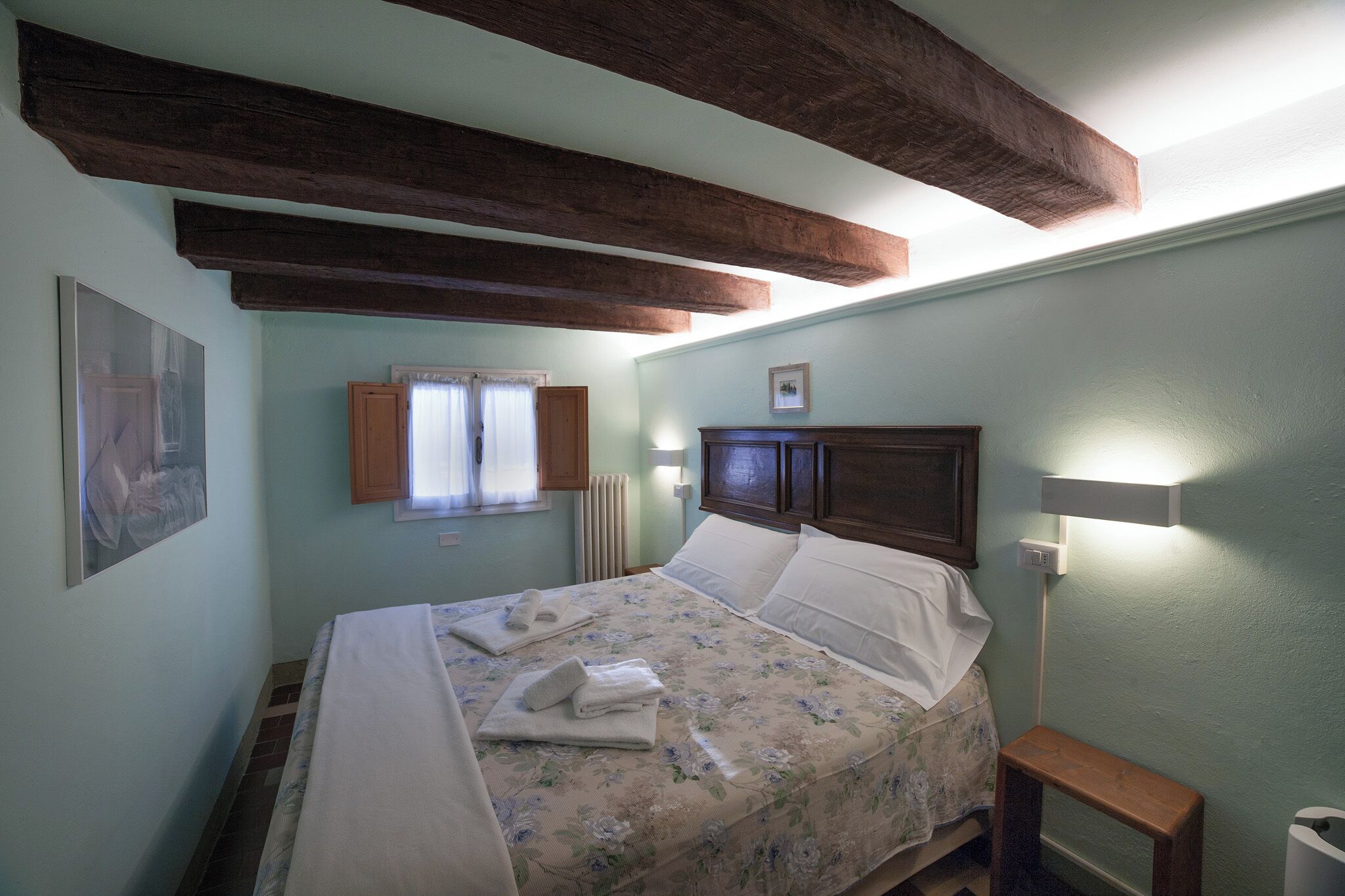 A bedroom with a bed, wooden beams, a nightstand, a wall-mounted light, and a framed picture.