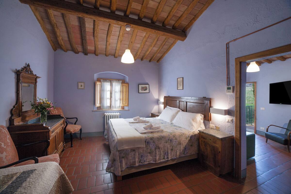 A bedroom with a wooden ceiling, a bed with a floral bedspread, a dresser, a mirror, and a window with curtains.