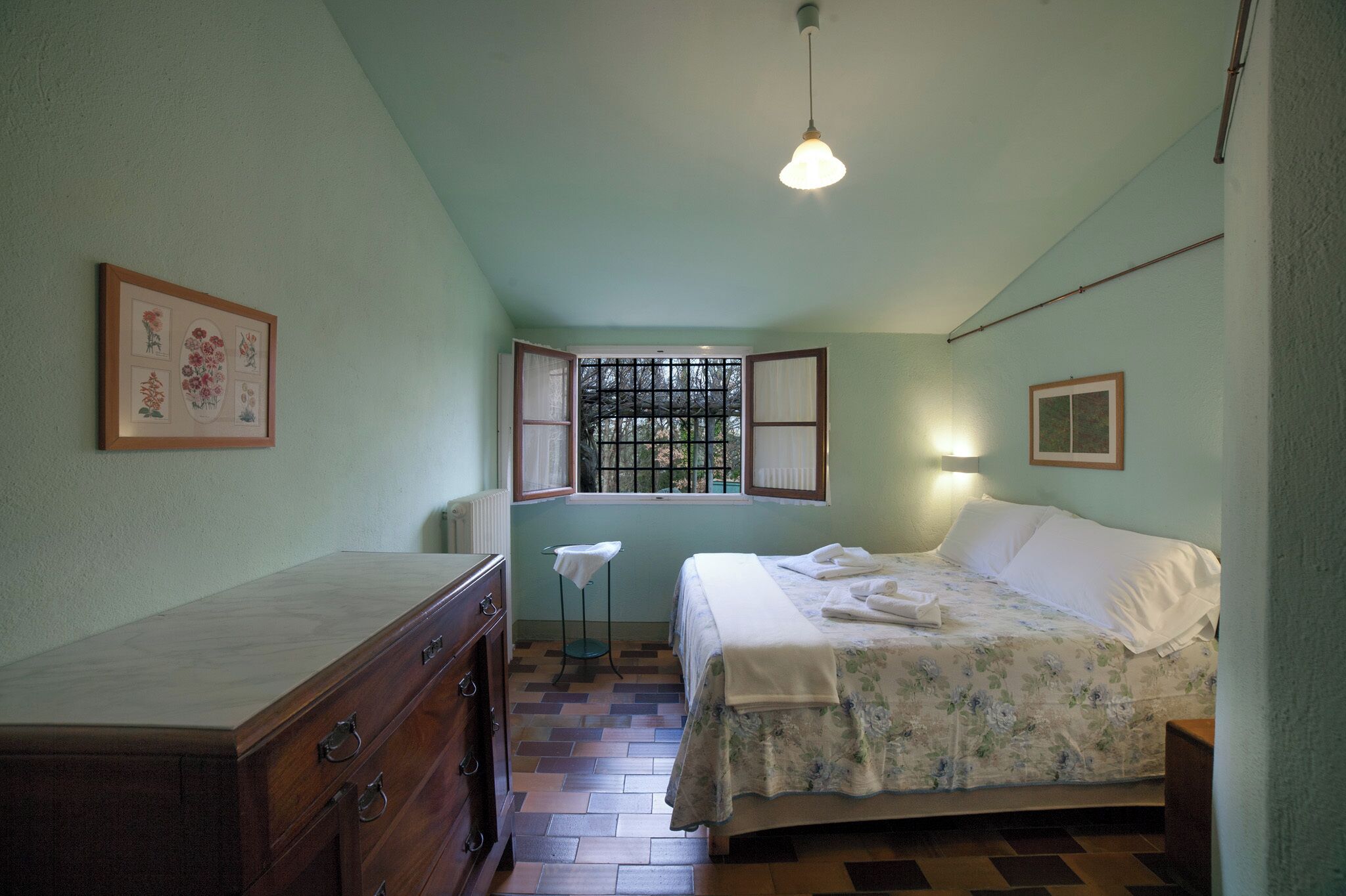 A bedroom with a bed, a dresser, a window with bars, and a hanging light fixture.