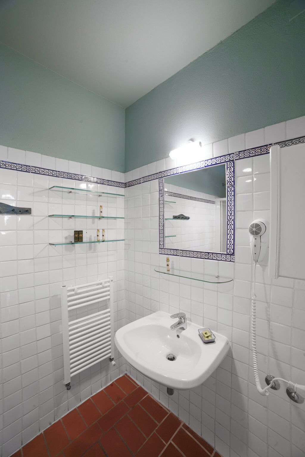 Une salle de bain avec des murs recouverts de carreaux blancs, un lavabo blanc, une étagère en verre, un miroir et une douche.