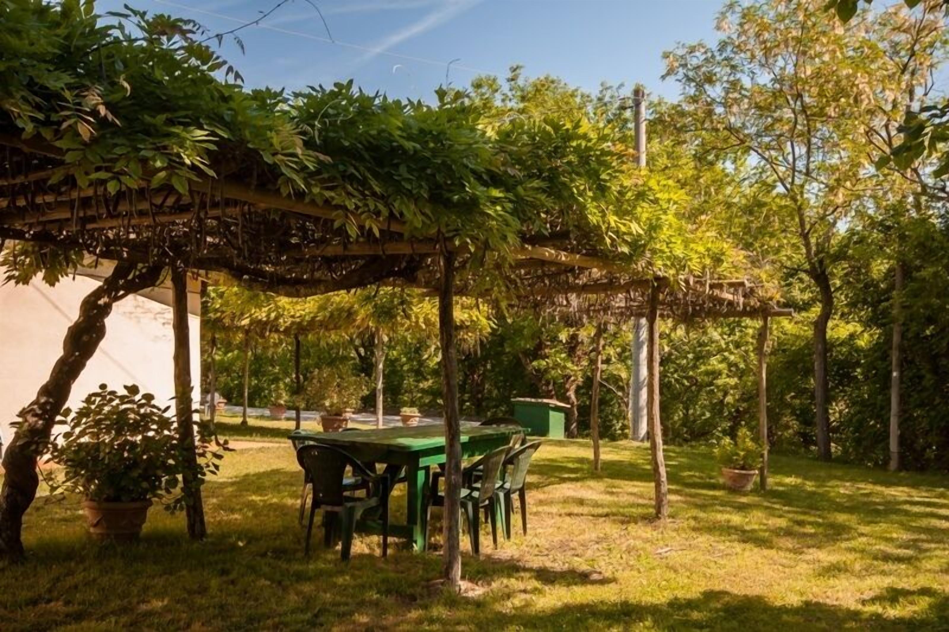 A covered outdoor seating area with a table and chairs, surrounded by trees and greenery.
